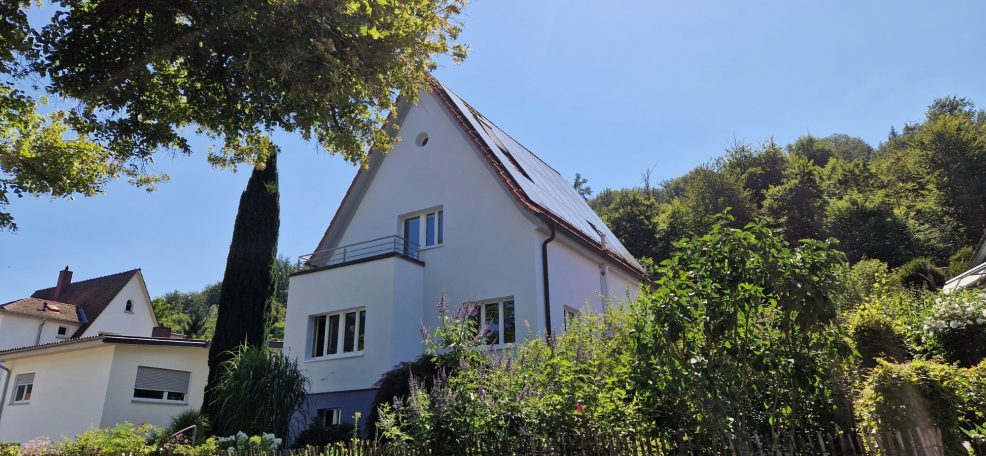 Fassadengestaltung Helles Haus mit Garten, umgeben von Bäumen und Hügeln unter blauem Himmel.