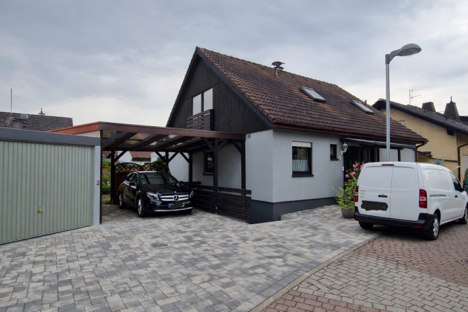 Fassadengestaltung Ein Einfamilienhaus mit Carport und Fahrzeugen auf einem gepflasterten Grundstück.